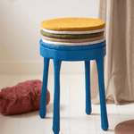 Colorful felt wool seat cushions stacked on a blue stool in a cozy indoor setting.