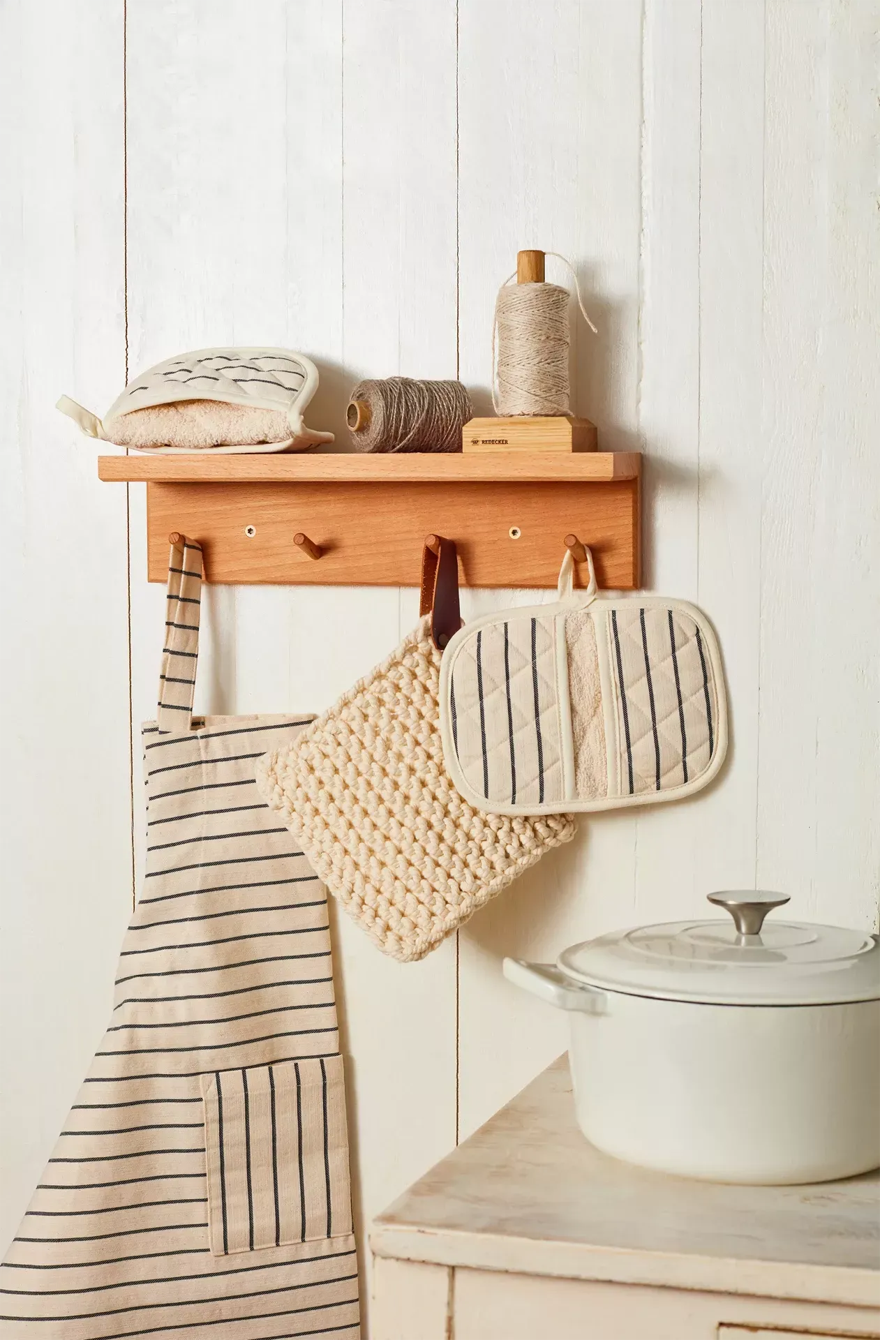 Wooden shelf with kitchen items including an apron, pot holders, and a white pot on a light wooden surface.