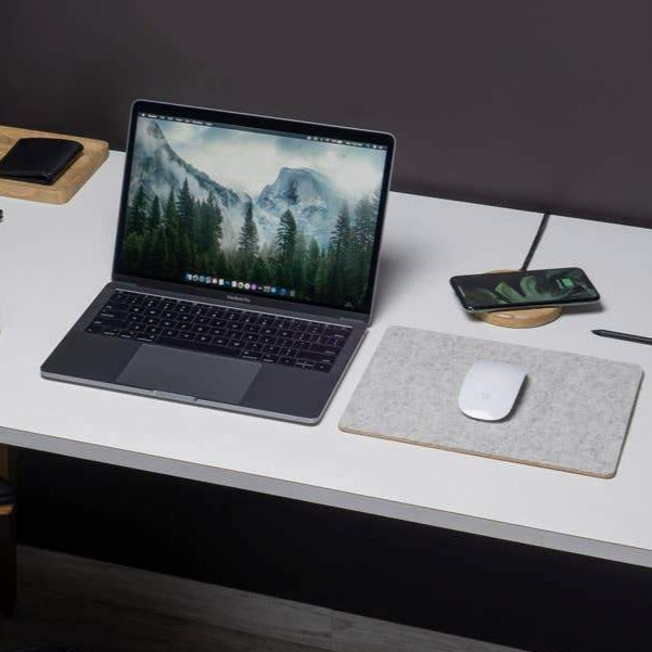 Desktop with open laptop, a felt mousepad with a mouse on it next to the laptop, and a phone charging on a wood wireless charger. 