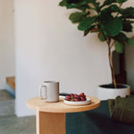 15 oz Hasami Porcelain mug in gloss grey is shown resting on a wood side table with a round top, next to a small white plate of purple grapes. There is a large ficus tree in the background against a white wall, and the corner of a dark green sofa is shown next to the side table.