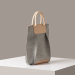 A Frankie Merino Wool Felt Tote in a grey color with beige leather accents on a white base displayed in a side-view
