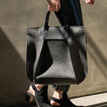 A charcoal-colored Frankie Merino Wool Felt Tote held by the top handle on a woman's side while walking, highlighting its chic design