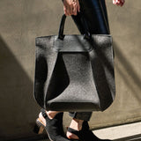 A charcoal-colored Frankie Merino Wool Felt Tote held by the top handle on a woman's side while walking, highlighting its chic design