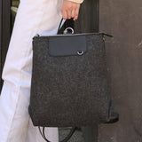 A charcoal-colored Bedford Merino Wool Felt Backpack held by the top handle on a woman's side while standing, highlighting its chic design