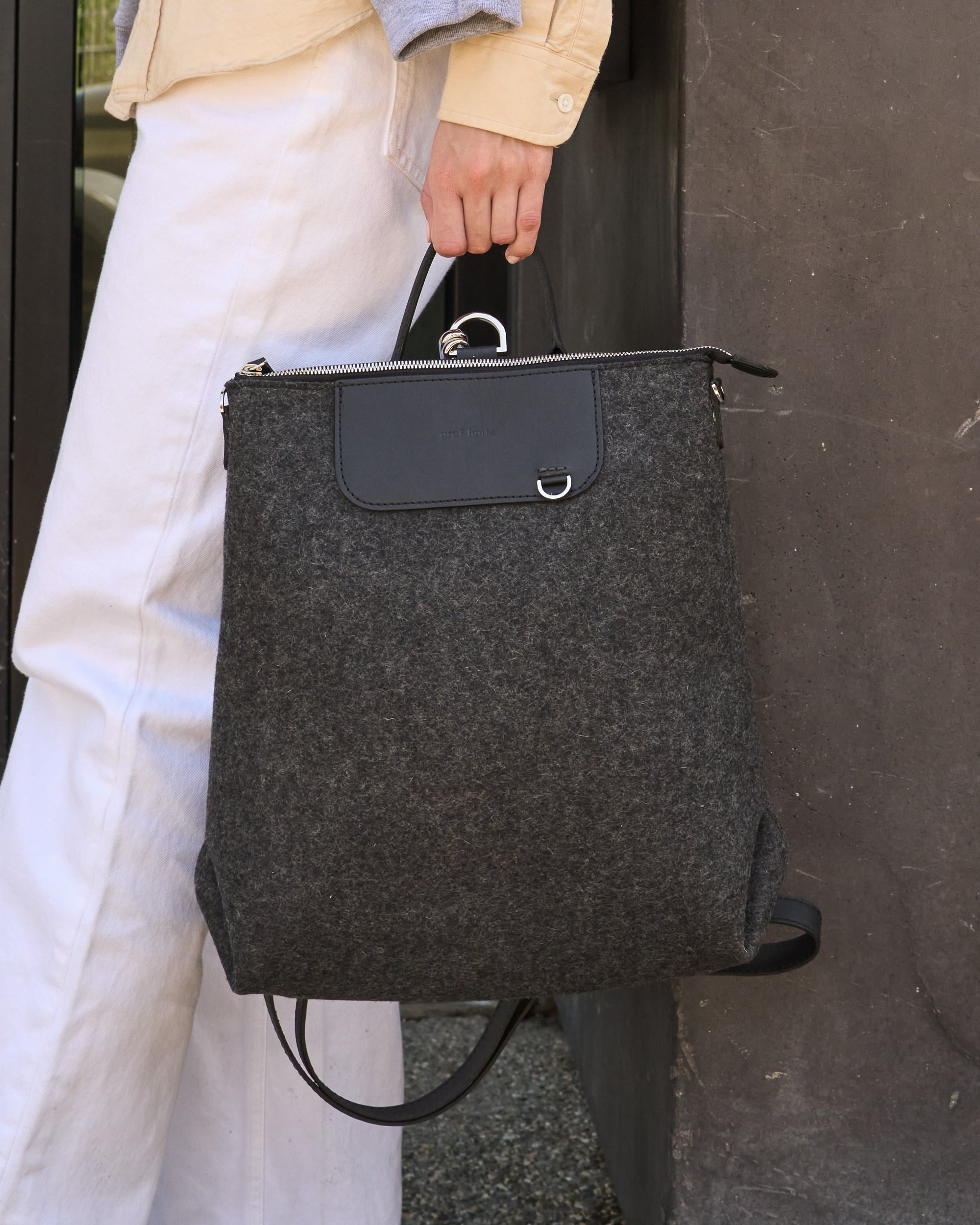 A charcoal-colored Bedford Merino Wool Felt Backpack held by the top handle on a woman's side while standing, highlighting its chic design