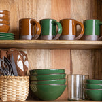 Wooden shelf with stacked glossy green bowls, clear glasses, utensils in a basket, and glossy brown and green ceramic mugs.