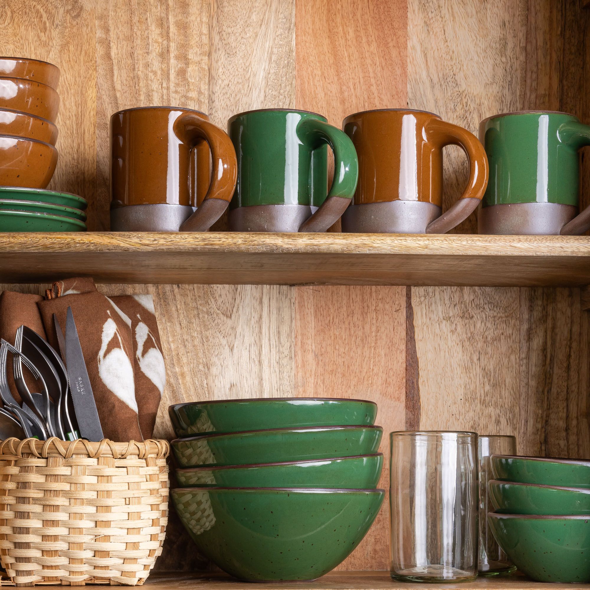 Wooden shelf with stacked glossy green bowls, clear glasses, utensils in a basket, and glossy brown and green ceramic mugs.