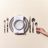 A hand arranging marble-handled flatware around a stack of cool white ceramic plates and a white dinner napkin.