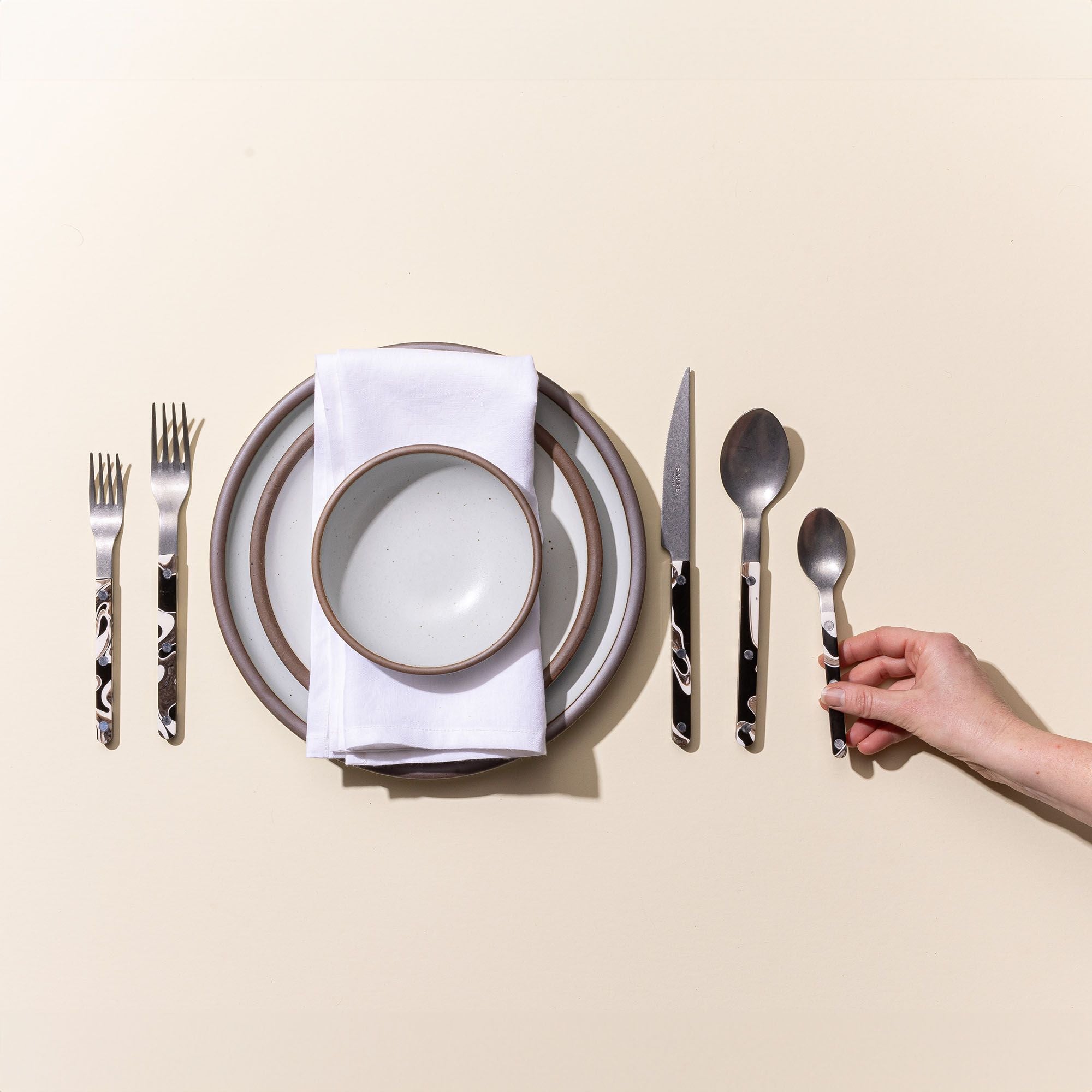 A hand arranging marble-handled flatware around a stack of cool white ceramic plates and a white dinner napkin.
