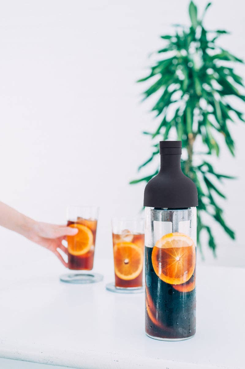 Bottle of dark liquid with orange slices, two glasses with similar contents, and a plant in the background.