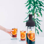 Bottle of dark liquid with orange slices, two glasses with similar contents, and a plant in the background.