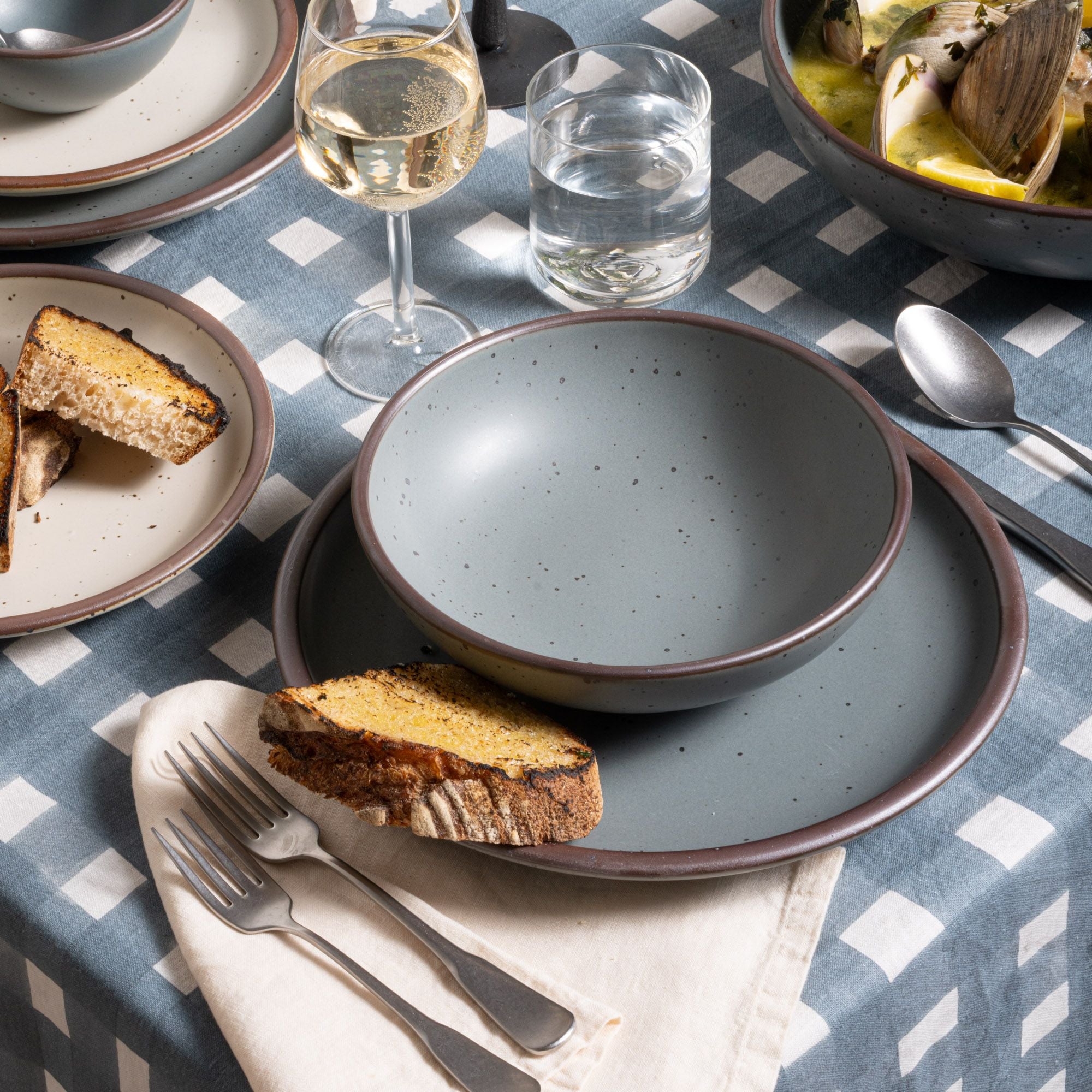 Medium shallow bowl and dinner plate in a blue-grey color, with silverware on a blue-grey crosshatch tablecloth