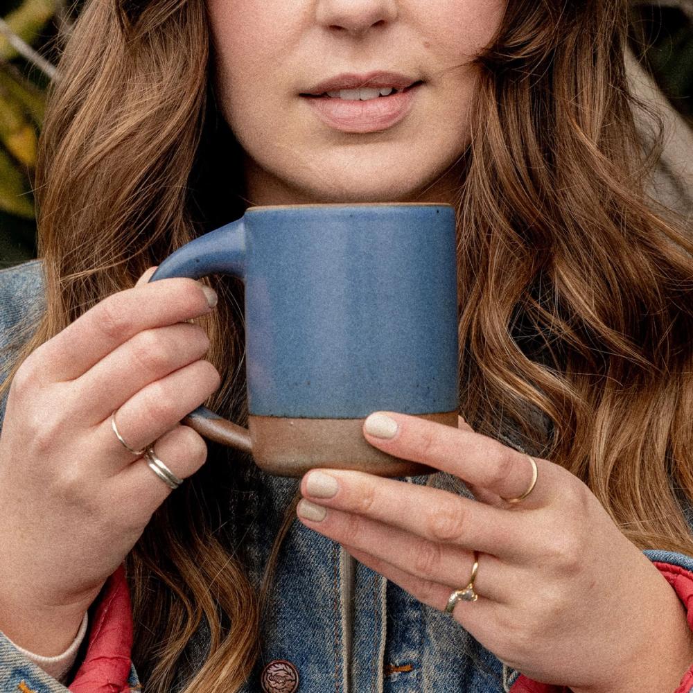 A woman holding The Mug in a cool medium blue color with two hands near her face outside.