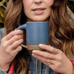 A woman holding The Mug in a cool medium blue color with two hands near her face outside.