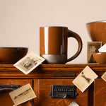 Glossy caramel brown ceramic mug and bowls on a wooden cabinet with vintage black-and-white photographs.