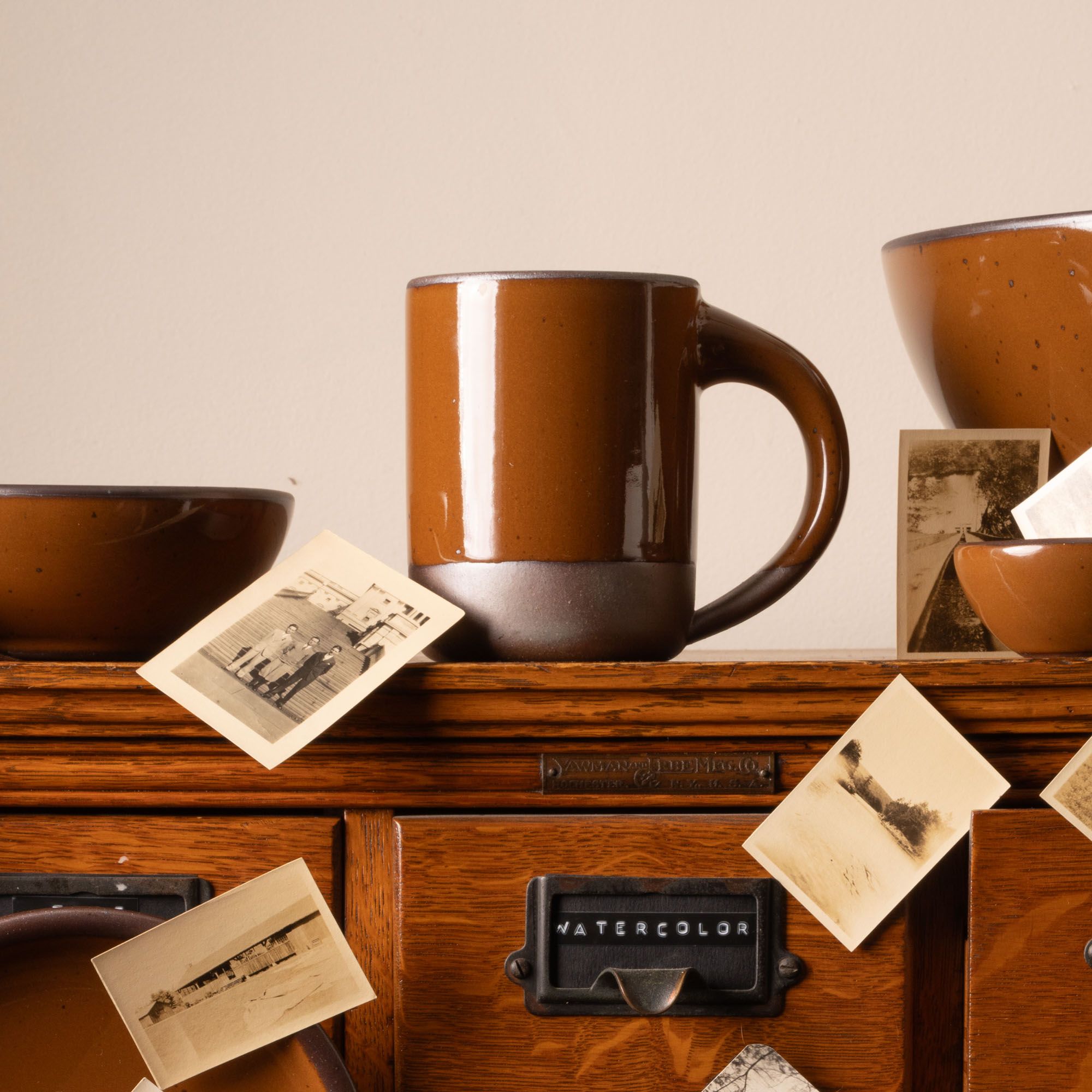 Glossy caramel brown ceramic mug and bowls on a wooden cabinet with vintage black-and-white photographs.