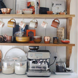 A photo by @lesleywgraham. A cozy coffee nook with hanging mugs, stacked dishes, and a silver espresso machine surrounded by jars of beans and sugar.