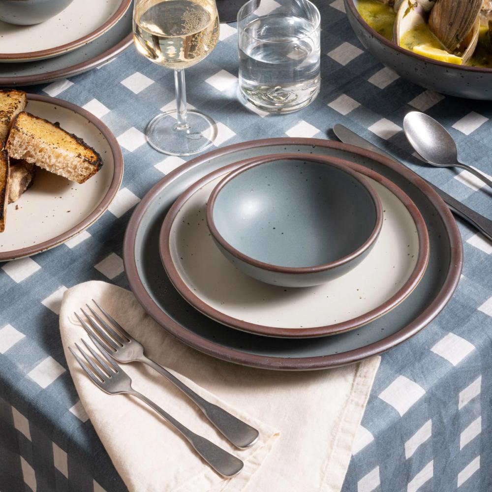 Small shallow bowl and dinner plate in a blue-grey color with a cream side plate, with silverware on a blue-grey crosshatch tablecloth