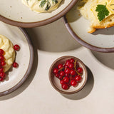 A tiny ceramic bowl in an off white color with little red berries inside sits next to plates of different sizes in the same color.