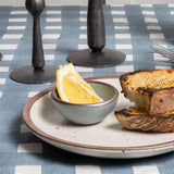 A tiny round bowl in a blue-grey color with a slice of lemon inside, sitting on a cream plate with toast.