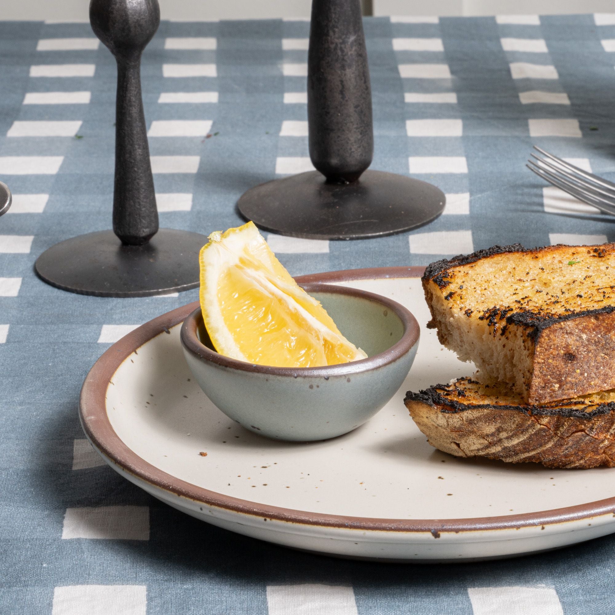 A tiny round bowl in a blue-grey color with a slice of lemon inside, sitting on a cream plate with toast.