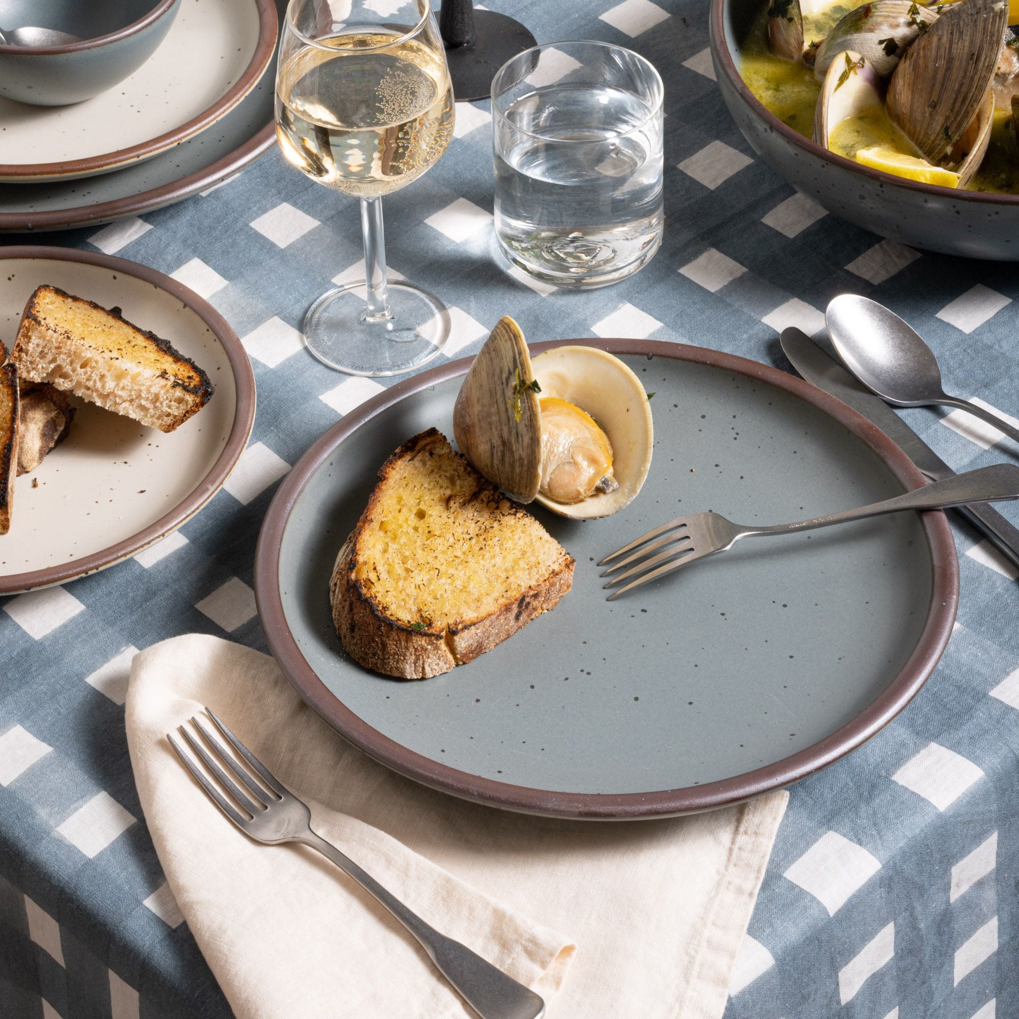 Clams and toast on a blue-grey dinner plate with silverware on a blue-grey crosshatch tablecloth