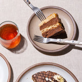 A small slice of cake on a ceramic cake plate in a pale light brown color with sophisticated forks and a glass of red juice.