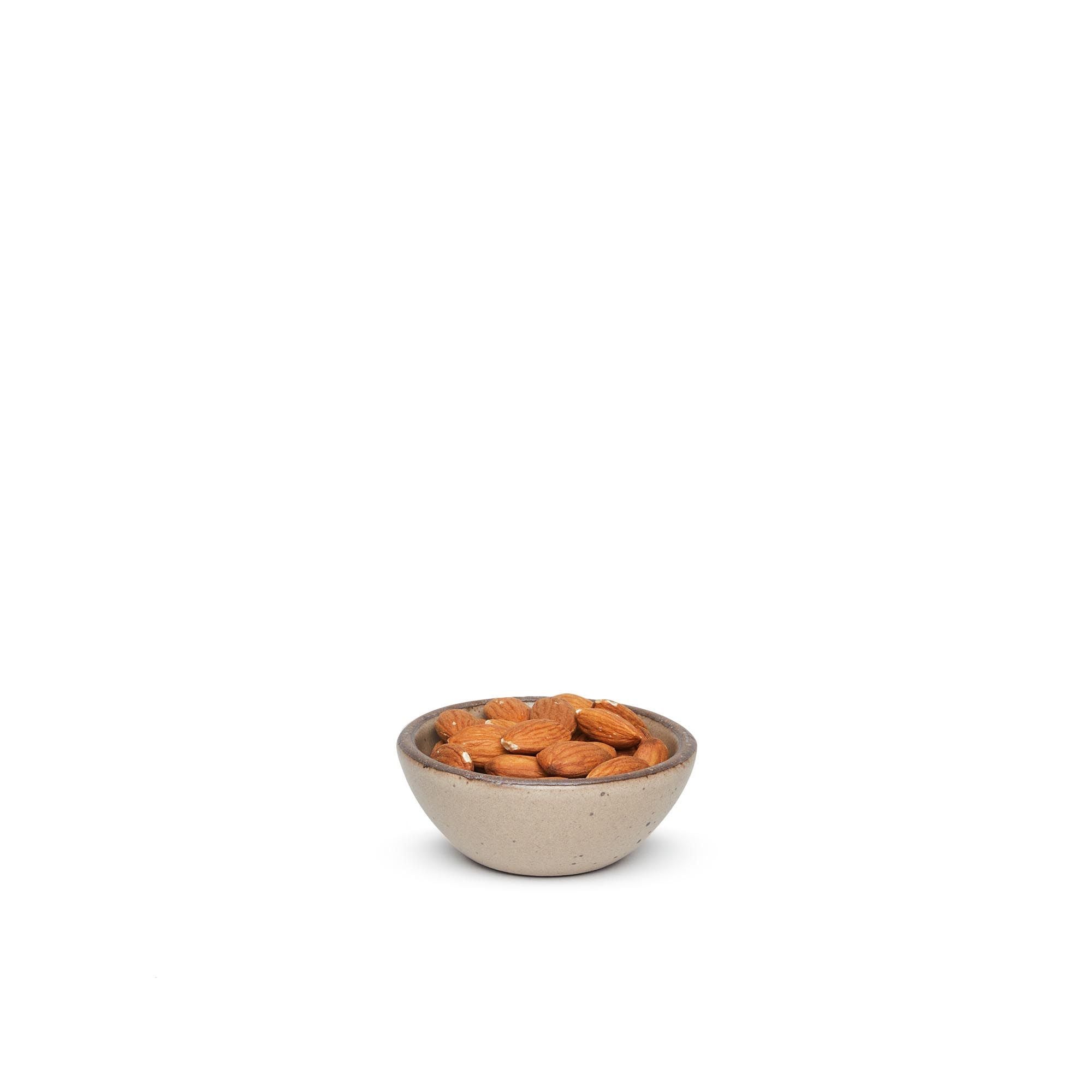 A tiny rounded ceramic bowl in a warm pale brown color featuring iron speckles and an unglazed rim, filled with almonds