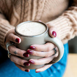 Photo by @farmettemichigan. Hands wrapping around a medium sized ceramic mug with handle in a warm pale brown color featuring iron speckles.