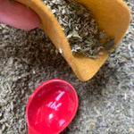 Yellow scoop filled with dried herbs on a textured surface with a red container labeled '1 tbl' in the foreground.
