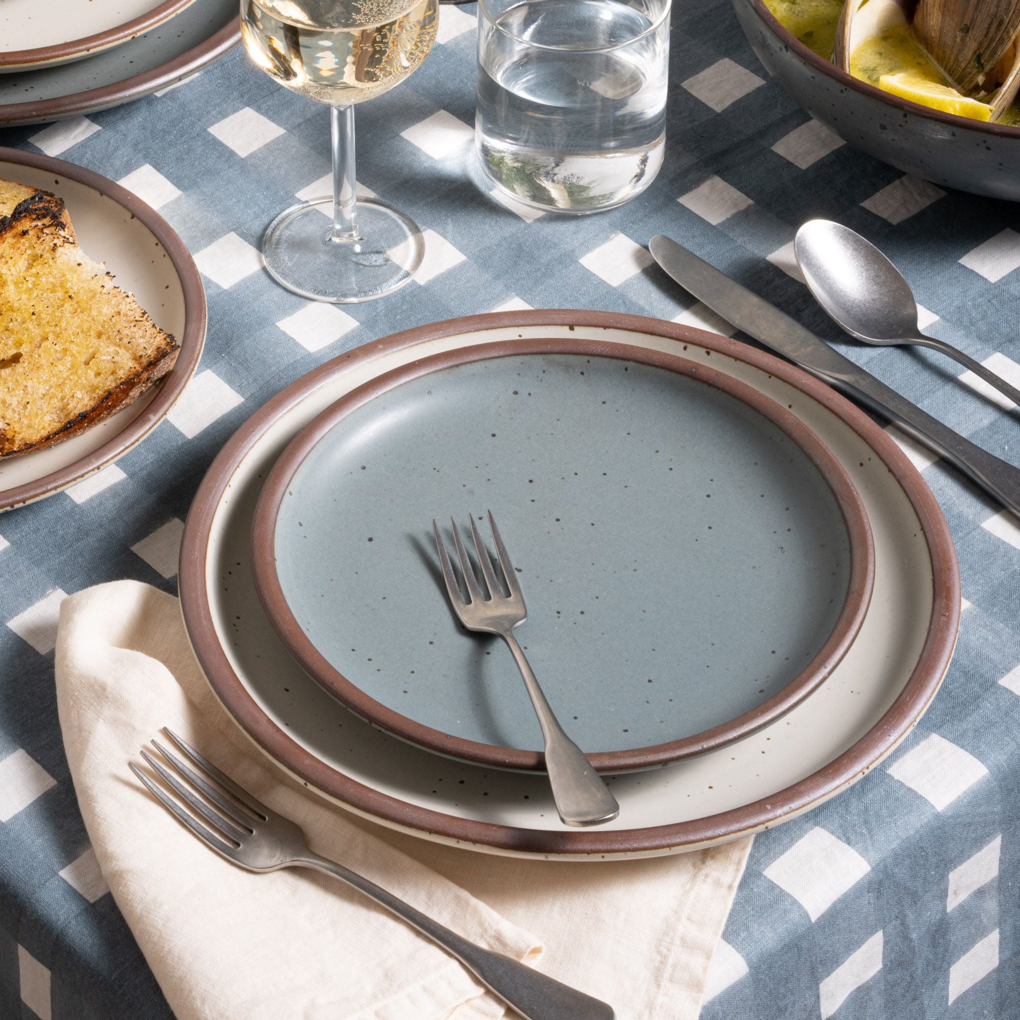 A place setting featuring a side blue-grey plate, large cream dinner plate, with silverware on a blue crosshatch tablecloth.