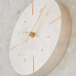 A minimalist round wall clock with a silver face shown in profile, featuring slim gold-tone hands and hour markers, mounted on a textured concrete wall.
