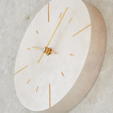 A minimalist round wall clock with a silver face shown in profile, featuring slim gold-tone hands and hour markers, mounted on a textured concrete wall.