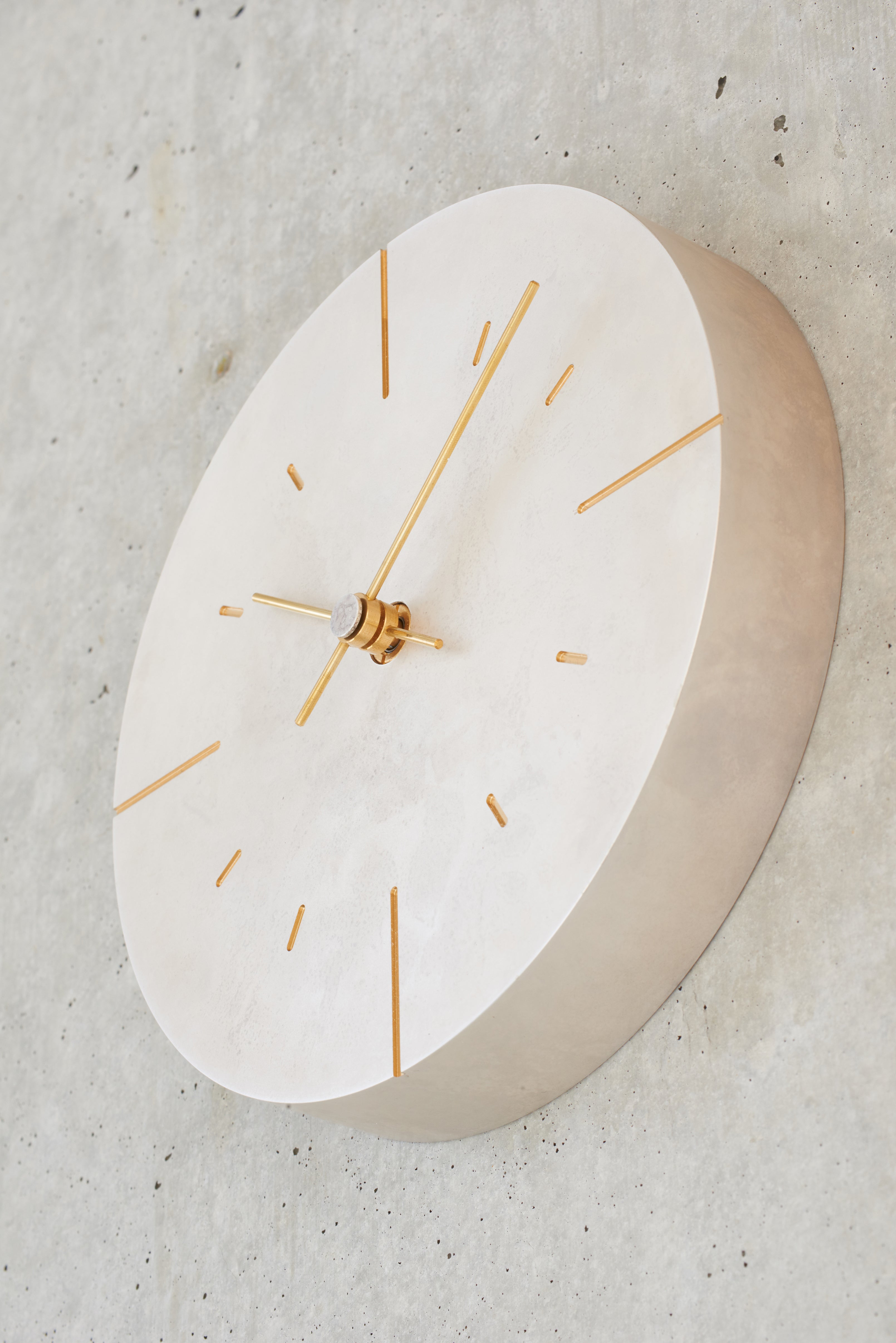 A minimalist round wall clock with a silver face shown in profile, featuring slim gold-tone hands and hour markers, mounted on a textured concrete wall.