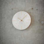 A minimalist round wall clock with a light silver face and gold-toned hands and hour markers, mounted on a textured gray concrete wall.