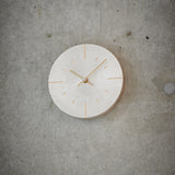 A minimalist round wall clock with a light silver face and gold-toned hands and hour markers, mounted on a textured gray concrete wall.