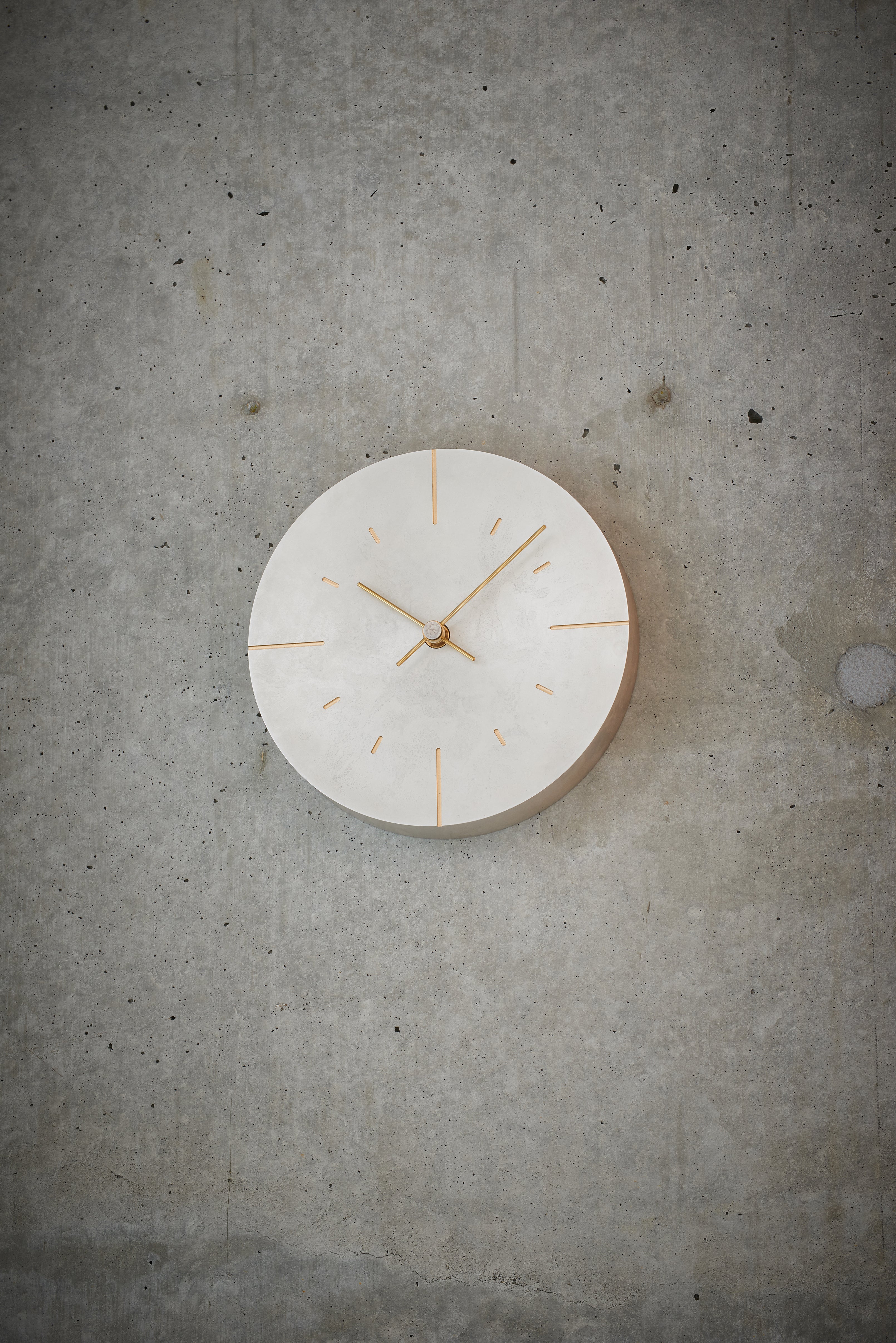 A minimalist round wall clock with a light silver face and gold-toned hands and hour markers, mounted on a textured gray concrete wall.
