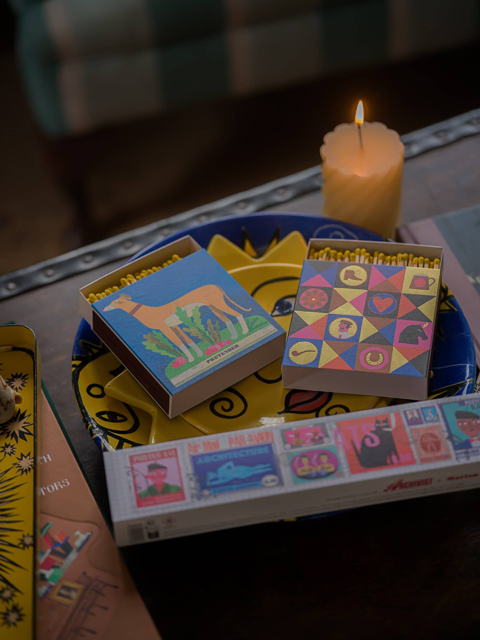 Matches boxes with colorful designs on a table with a lit candle in the background