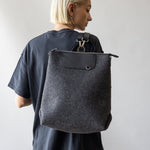 A person with short blond hair, in a dark t-shirt, is turned sideways holding a large graf lantz Merino Wool Backpack. Known for its sustainability, this bag combines durability and style against a plain, light background.
