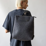 A person with short blond hair, in a dark t-shirt, is turned sideways holding a large graf lantz Merino Wool Backpack. Known for its sustainability, this bag combines durability and style against a plain, light background.