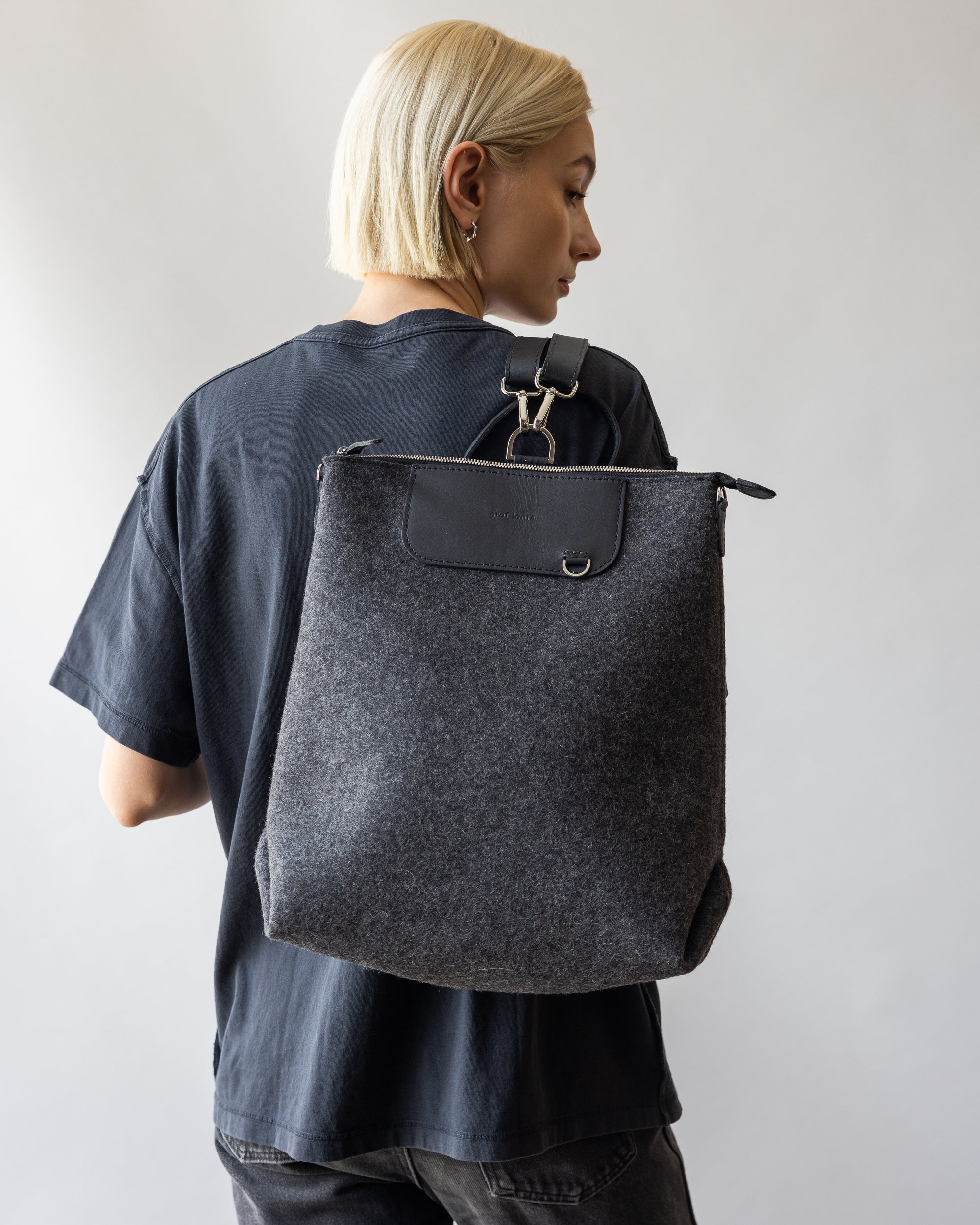 A person with short blond hair, in a dark t-shirt, is turned sideways holding a large graf lantz Merino Wool Backpack. Known for its sustainability, this bag combines durability and style against a plain, light background.