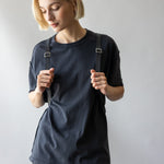 A person with short blonde hair, wearing a black t-shirt and dark jeans, stands against a plain background. They hold the straps of their graf lantz Merino Wool Backpack on their shoulders and gaze down with a neutral expression, showcasing its blend of durability and style.