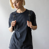 A person with short blonde hair, wearing a black t-shirt and dark jeans, stands against a plain background. They hold the straps of their graf lantz Merino Wool Backpack on their shoulders and gaze down with a neutral expression, showcasing its blend of durability and style.