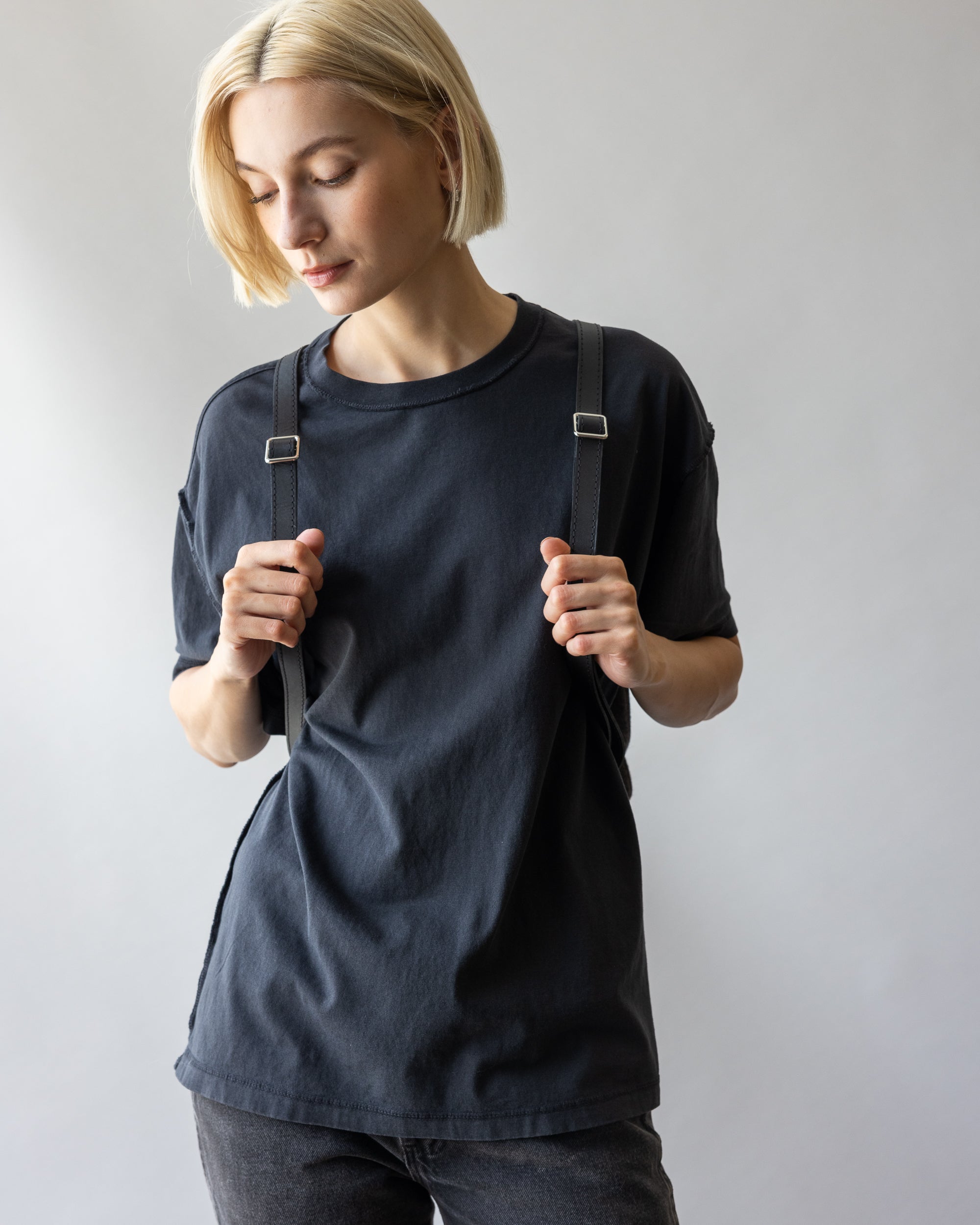 A person with short blonde hair, wearing a black t-shirt and dark jeans, stands against a plain background. They hold the straps of their graf lantz Merino Wool Backpack on their shoulders and gaze down with a neutral expression, showcasing its blend of durability and style.