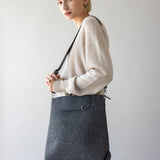 A person with short blond hair is wearing a light beige sweater, holding a large gray Merino Wool Backpack by graf lantz, renowned for its durability and style. The softly lit background is plain.
