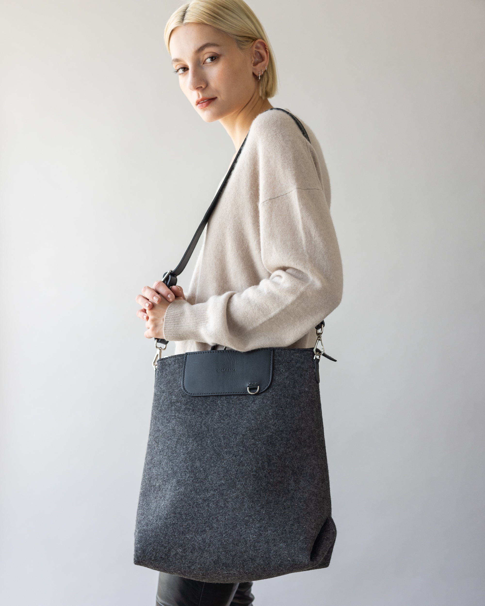 A person with short blond hair is wearing a light beige sweater, holding a large gray Merino Wool Backpack by graf lantz, renowned for its durability and style. The softly lit background is plain.