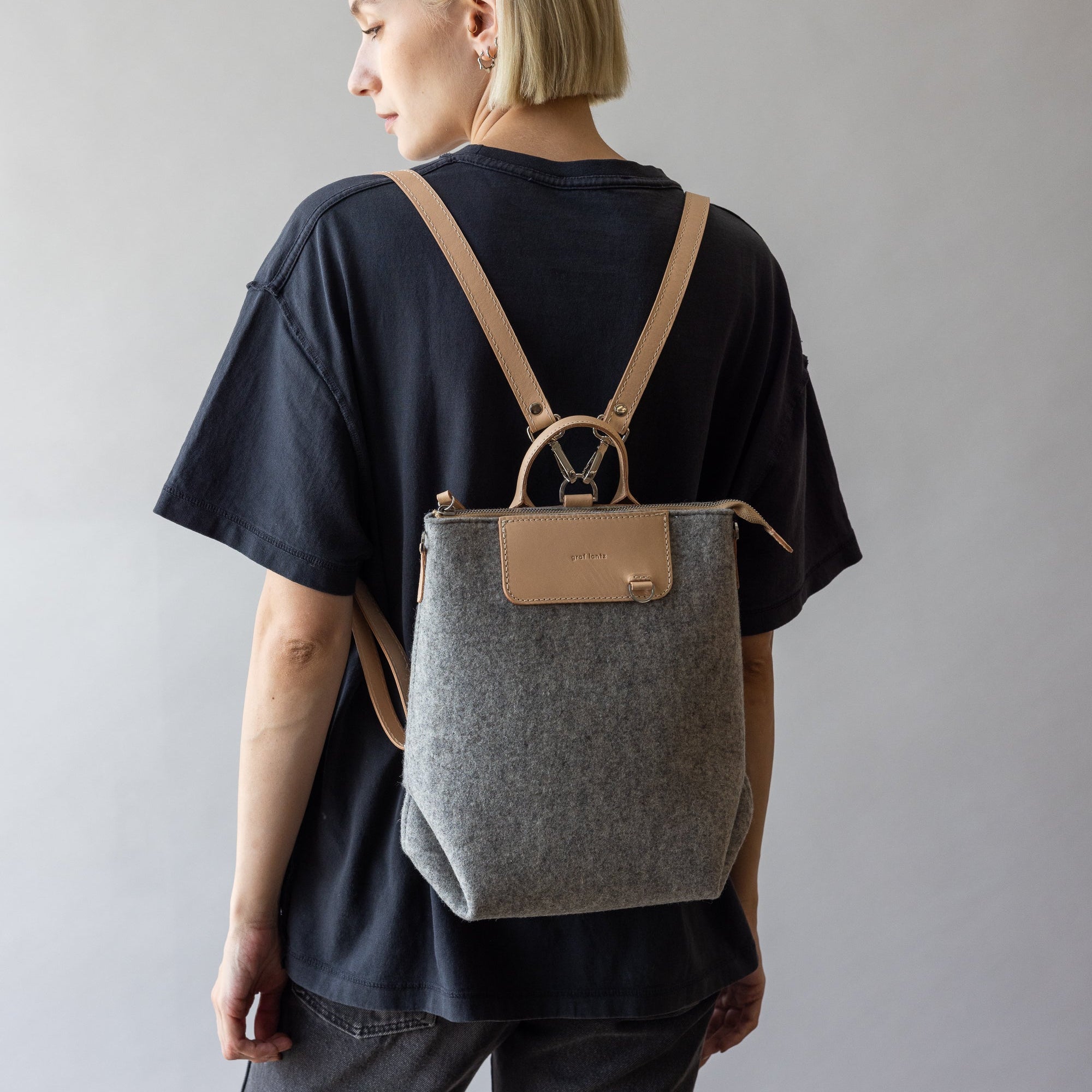 A person with short blonde hair, in a black t-shirt and jeans, faces away while carrying a versatile graf lantz backpack in gray and beige with leather straps. The multifunctional bag adds flair against the neutral background.