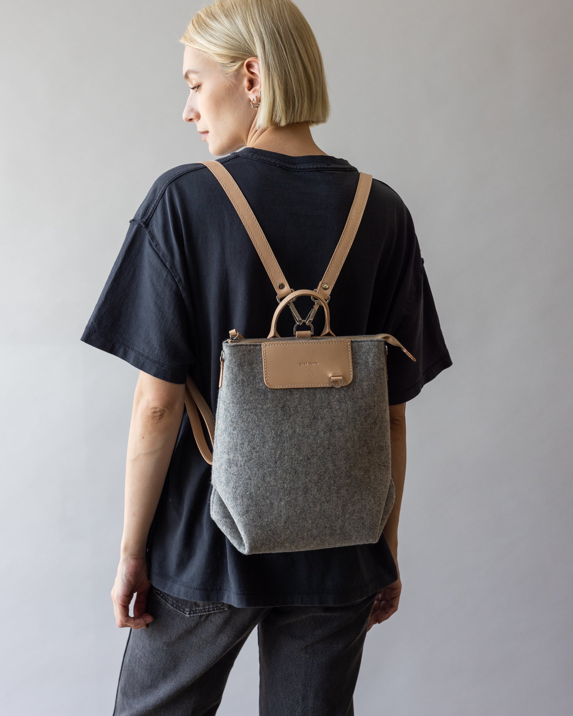 A person with short blonde hair, in a black t-shirt and jeans, faces away while carrying a versatile graf lantz backpack in gray and beige with leather straps. The multifunctional bag adds flair against the neutral background.