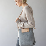 A person with short blonde hair wears a beige sweater and black pants, holding a graf lantz shoulder bag. They face sideways against a neutral background, highlighting the bags versatile design.