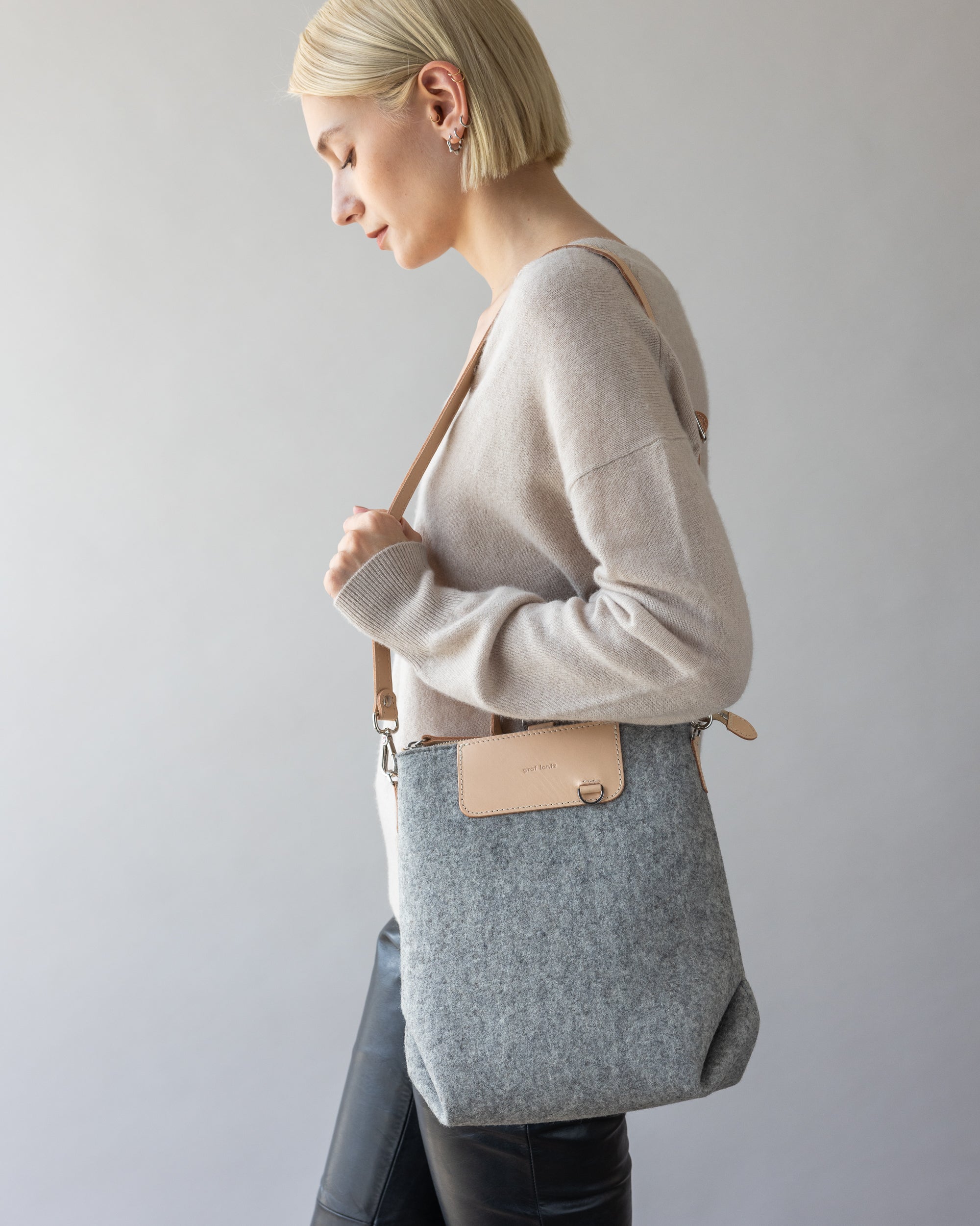 A person with short blonde hair wears a beige sweater and black pants, holding a graf lantz shoulder bag. They face sideways against a neutral background, highlighting the bags versatile design.